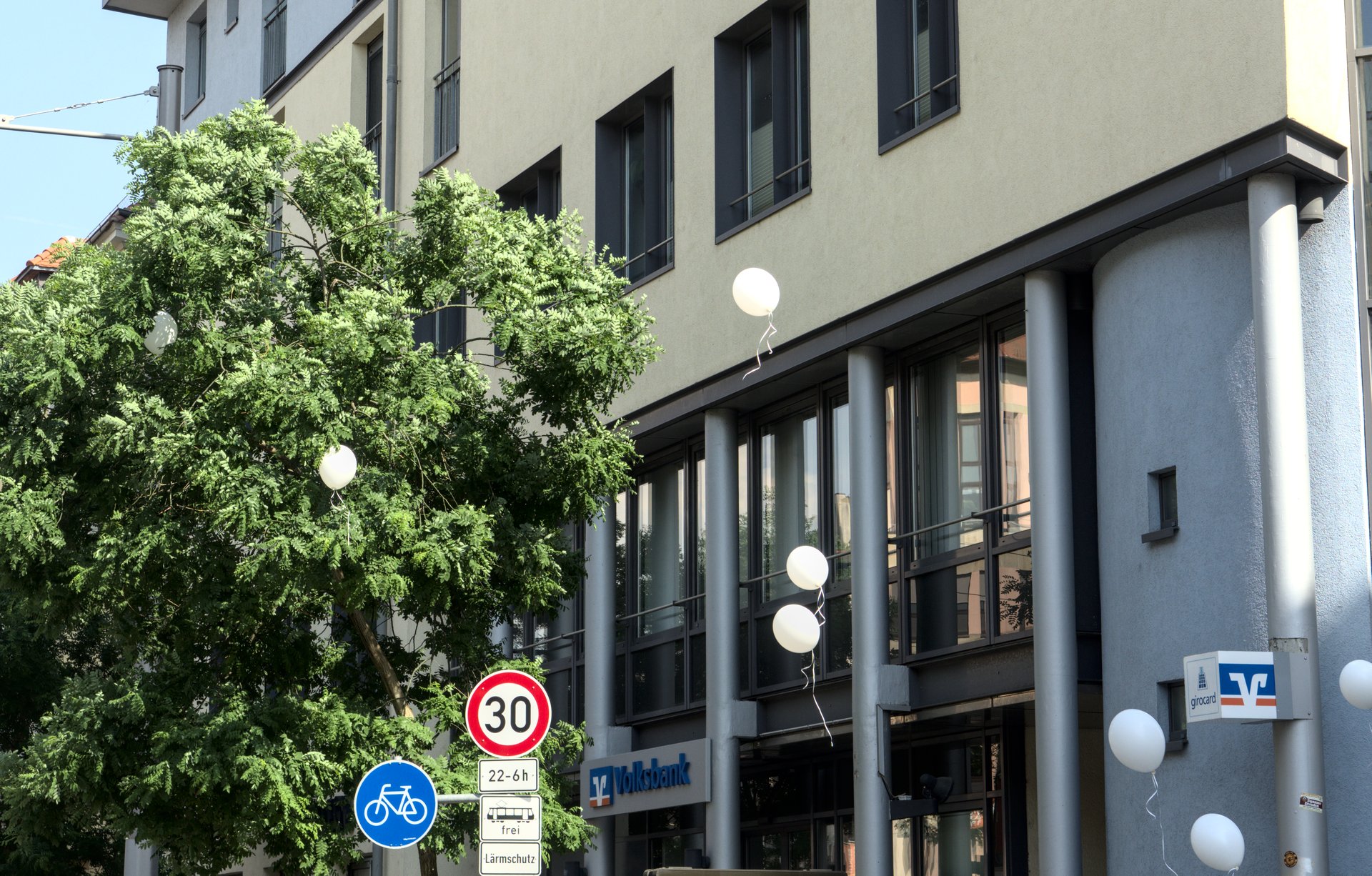 aufsteigende weiße Luftballons vor einer Hausfassade, links das Verkehrsschild an dem Ghostbike #16 angeschlossen ist